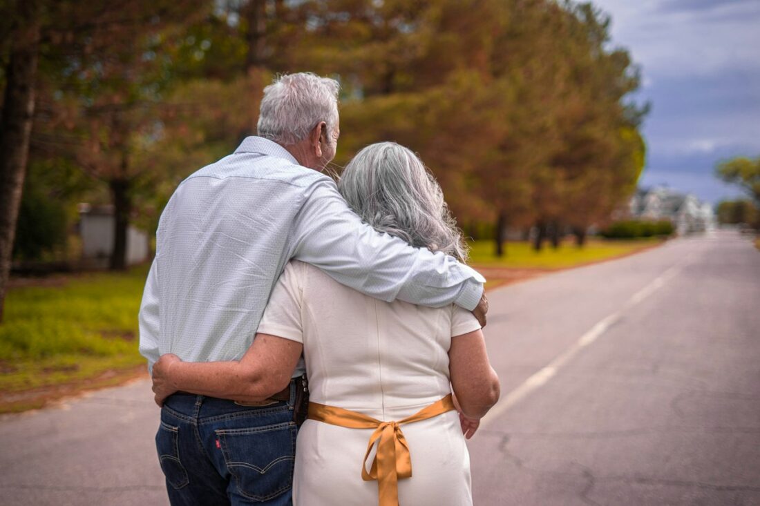 Casal de cabelos grisalhos caminhando abraçado em uma estrada arborizada