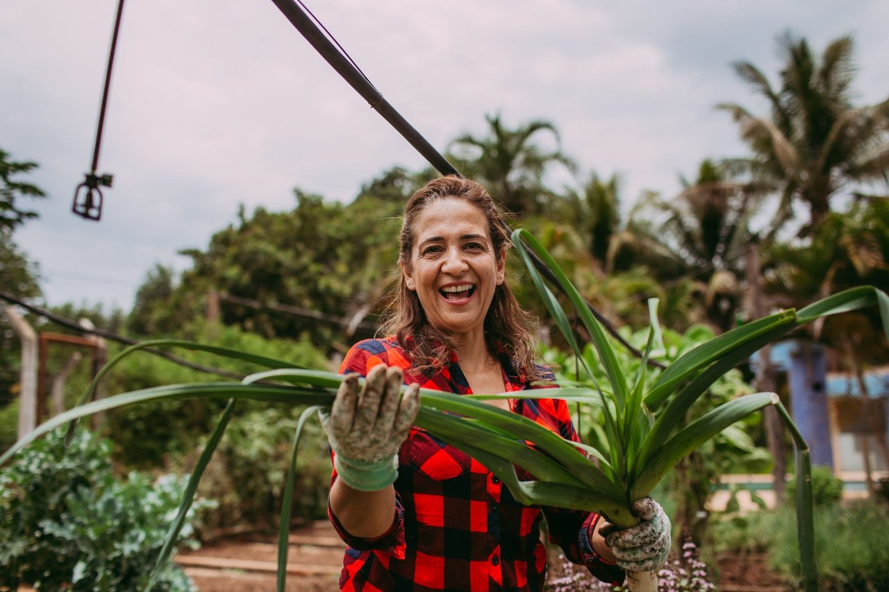 Mulher de camisa xadrez vermelha sorrindo enquanto segura uma planta no quintal
